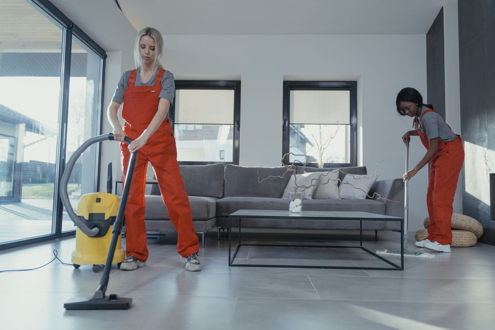 Two cleaners in orange overalls vacuuming and mopping a bright living room