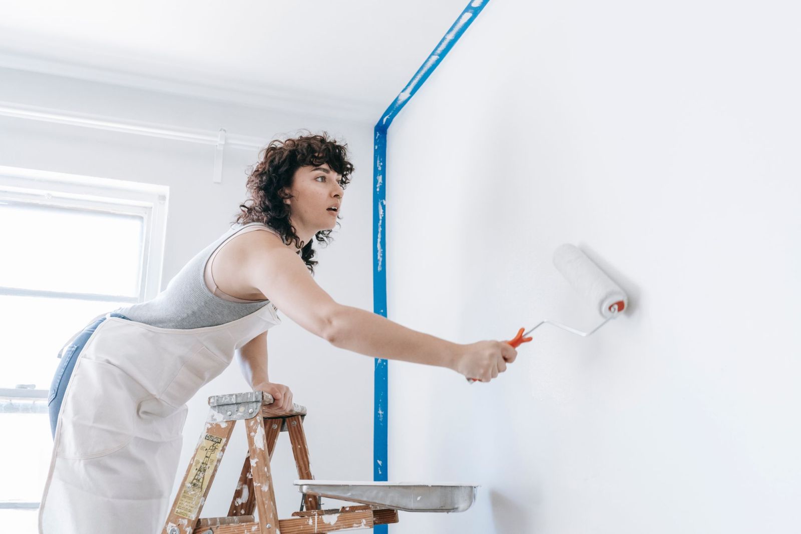 A decorator on a step ladder rolling white paint onto a wall